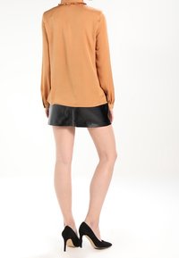 Femme debout portant un chemisier beige à manches longues, une mini-jupe en cuir noire et des chaussures à talons noirs, de dos à l'appareil photo.