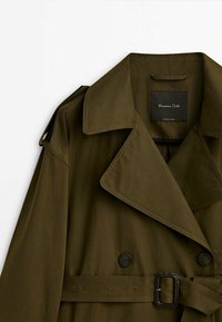 Oliwkowy płaszcz trencherowy z dużym kołnierzem, podwójnymi guzikami i paskiem. Posiada pagon na ramieniu i gładką fakturę.