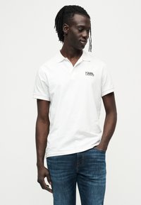 Λευκό πόλο πουκάμισο με κοντά μανίκια, με μαύρο λογότυπο Karl Lagerfeld. Φορεμένο με σκούρο μπλε τζιν. Απλός σχεδιασμός, ελαφρύ ύφασμα.