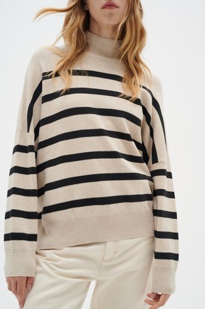 Maglione beige con righe orizzontali nere, colletto alto e maniche ampie. Il tessuto appare morbido e lavorato a maglia. Abbinato a pantaloni di colore chiaro.
