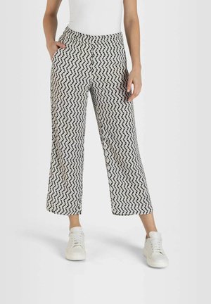 Person wearing black-and-white zigzag patterned wide-leg pants, white tank top, and white sneakers, standing with one hand in pocket.