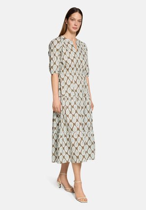 Frau trägt ein beige-blau gemustertes Midikleid mit Puffärmeln und beigefarbene, flache Sandalen mit Riemchen, steht vor einem schlichten Hintergrund.