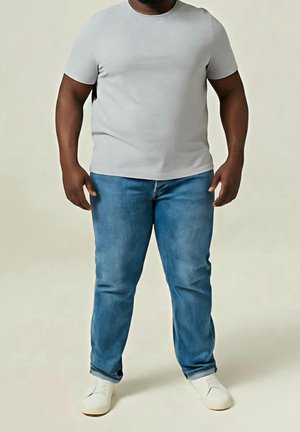 Homme debout, portant un t-shirt gris clair, un jean bleu et des chaussures blanches sans lacets, sur un fond neutre uni.