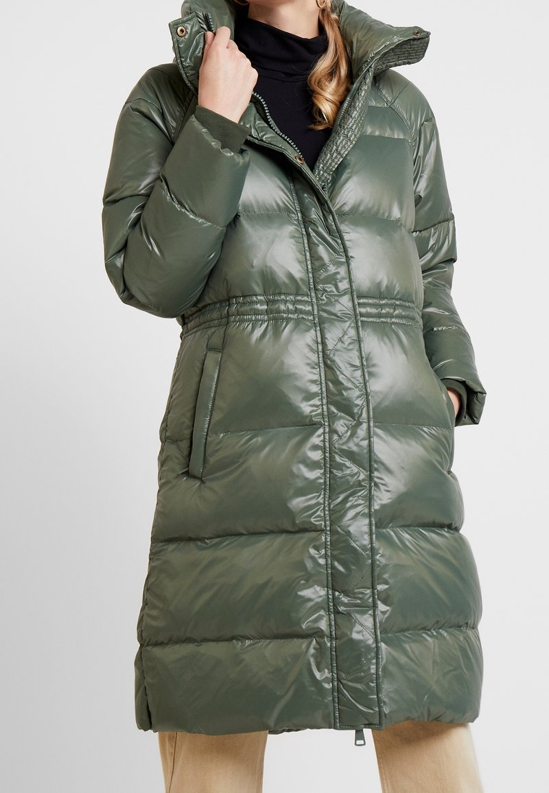 Olivinvihreä toppatakki kiiltävällä pinnalla, jossa on korkea kaulus, vyötäröltä kapea malli ja kaksi sivutaskua.