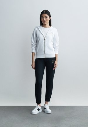 Mujer de pie con sudadera blanca con cremallera, camiseta blanca, pantalones deportivos negros y zapatillas blancas sobre un fondo liso gris claro.