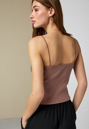 Junge Frau mit glatten braunen Haaren, die ein mauvefarbenes, geripptes Spaghettiträgertop und schwarze Hosen trägt, schaut über ihre linke Schulter nach links.