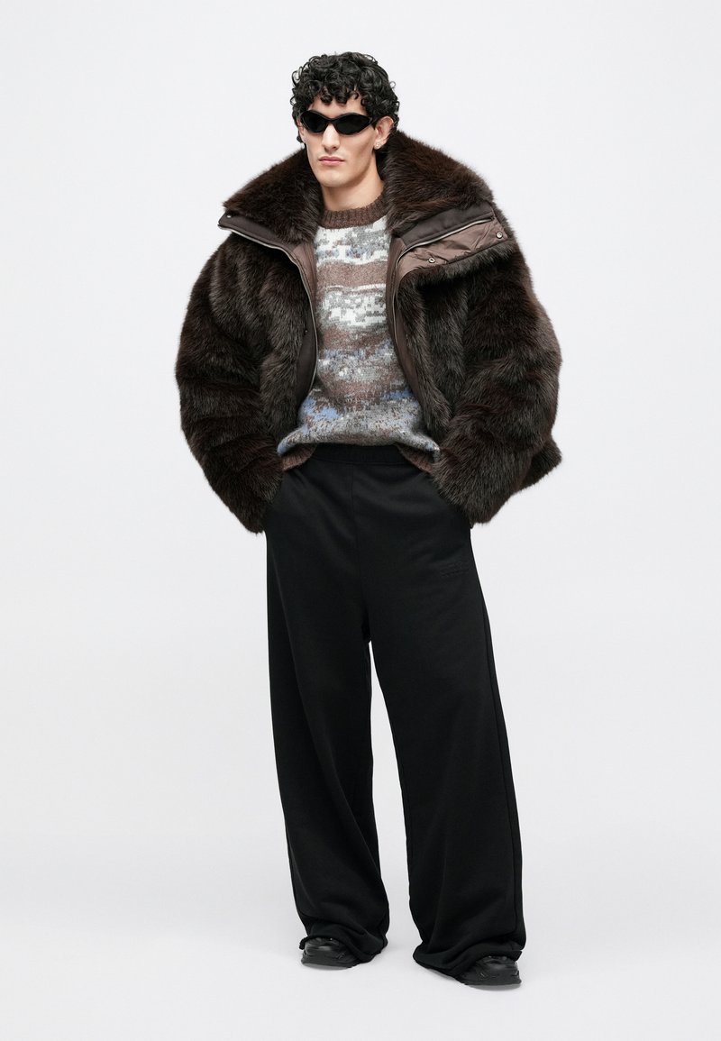 Brown faux fur jacket with a padded collar, knit sweater with gray and blue patterns, black wide-leg pants, and round dark sunglasses.