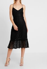 Svart spetsklänning med justerbara spaghetti-band, en figurnära bodice och en utställd fåll. Har transparenta lager av volanger i botten.