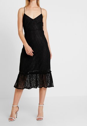 Musta pitsimekko, jossa on säädettävät spaghetti-hihnat, tiukka yläosa ja levenevä helma. Alhaalla on läpinäkyviä kerroksittaisia rypytyksiä.