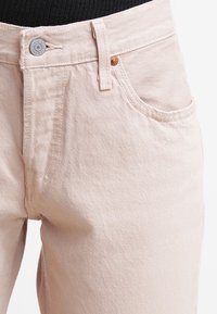 Pantalones de mezclilla beige claro con detalles en la cintura y parte superior del muslo, con bolsillo frontal, pasador para cinturón y cierre de botón metálico, llevados con una blusa negra.