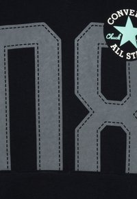 Fekete póló, amelyen nagy, szürke hímzett "08" számok és egy kör alakú, teal és fehér színű logó található, amely a "Converse Chuck All Star" feliratot viseli a tetején.