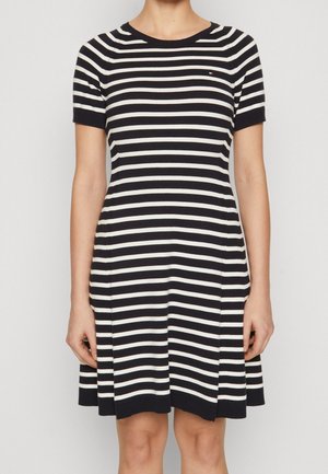 Femme portant une robe à manches courtes à rayures horizontales noires et blanches, avec un col rond, debout devant un fond uni.