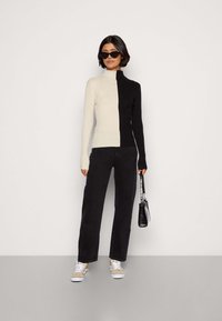 Rebrast turtleneck pulover v kremni in črni barvi, oprijetega kroja z dolgimi rokavi. V kombinaciji s črnimi hlačami in sproščenimi supergami, drži majhno torbico.