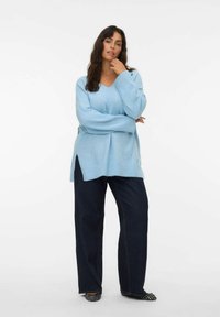 Lichtblauwe oversized trui met een V-hals, lange mouwen en zijsplitten, gecombineerd met donker wijde jeans en patroon flats.