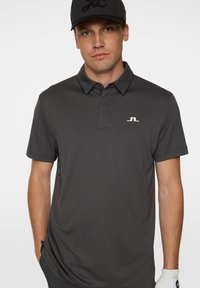 Kolgrå pikétröja med klassisk krage, korta ärmar och en diskret vit logotyp på bröstet. Slätt, lätt tyg.