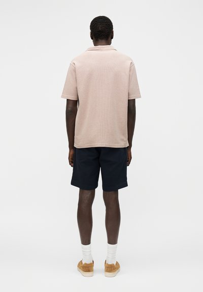 Homme portant un polo beige à manches courtes, un short foncé, des chaussettes blanches et des chaussures marron, debout face à un fond blanc uni.