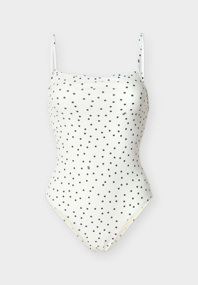 Λευκό ολόσωμο μαγιό με λεπτές τιράντες και μαύρες πουά διακοσμητικές κουκκίδες ομοιόμορφα κατανεμημένες στο ύφασμα, απεικονίζεται σε απλό φόντο.