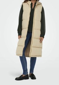 Beige gepolsterte lange Weste mit Kapuze, mit einem Reißverschluss und einem gesteppten Design. Kombiniert mit einer dunkelgrünen Bluse, blauen Jeans und schwarzen Mokassins.