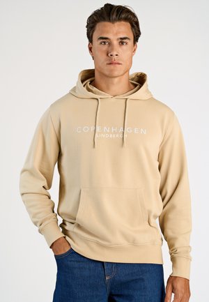 Beige hoodie met voorzak, capuchon met trekkoord en bedrukt met "COPENHAGEN" tekst. Zachte stof; gecombineerd met blauwe spijkerbroek.
