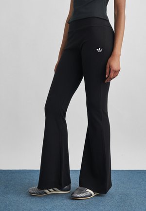 Vrouw draagt zwarte uitlopende broek met Adidas-logo op dij en zwarte Adidas slip-on schoenen, staand op een blauw tapijt.