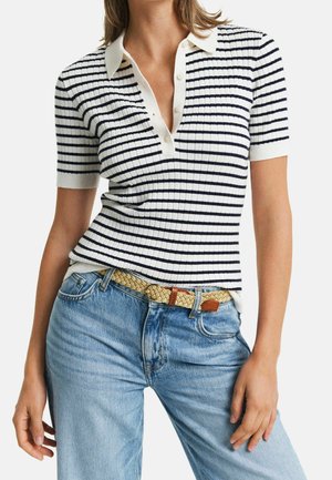 Vrouw die een witte polo met korte mouwen draagt met zwarte horizontale strepen en lichtblauwe spijkerbroek met een geweven beige riem.