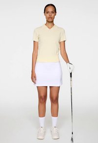 Vaaleankeltainen lyhythihainen puuvillapikapaita, yhdistetty valkoiseen golfhameeseen. Malli pitää kädessään golfmailaa ja hänellä on valkoiset sukat ja lenkkarit.