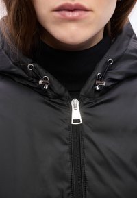 Κοντινό πλάνο ενός ατόμου που φοράει μια μαύρη φουσκωτή ζακέτα με ασημένιο φερμουάρ και κορδόνια για ρύθμιση πάνω σε μαύρο ζιβάγκο.