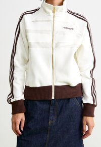 Vit zip-jacka med bruna detaljer, med ribbade muddar och nederkant, tre bruna ränder på ärmarna och en broderad logotyp.