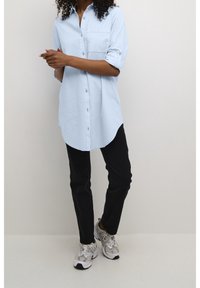 Chemise bleu clair à boutons avec manches retroussées, ourlet arrondi et poche poitrine, portée avec un pantalon noir et des chaussures de sport grises.