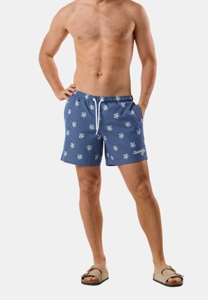 Oberkörperfreier Mann trägt blaue, gemusterte Badeshorts mit weißem Kordelzug und beigen Sandalen, steht mit den Händen auf der Hüfte und in den Taschen.