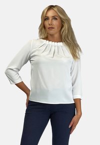 Biała bluzka z plisowanym dekoltem i rękawami trzyczwartowymi. Wykonana z gładkiej tkaniny, zestawiona z ciemnymi, strukturalnymi spodniami.