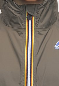 Oliwkowa kurtka z wysokim kołnierzem, z centralnym zamkiem błyskawicznym oraz żółtymi, białymi i granatowymi paskami. Logo umieszczone po lewej stronie.