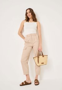 Beige, magas derekú cargo nadrág, oldalt zsebekkel, fehér tank toppal és barna szandálokkal párosítva. Egy textúrált szalma táska barna részletekkel a kezében.