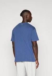 Modré bavlněné tričko s krátkými rukávy, kulatým výstřihem a malým logem na zadní straně. Zobrazeno ze zadního pohledu.