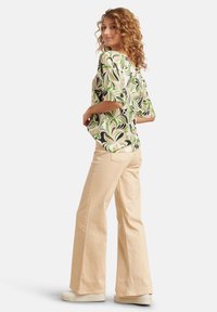 Bluse mit floralen Mustern in Grün, Schwarz und Creme; kombiniert mit beigen, weiten Hosen und weißen Schuhen. Glatte Stoffqualität, lockere Passform.