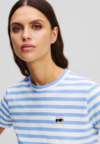 T-shirt rayé en coton bleu clair et blanc avec un col rond. Présente un petit design brodé sur la poitrine.
