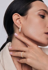 Złote kolczyki nałogowe z przezroczystymi cyrkoniami, z teksturowanym wykończeniem. Modelka nosi pasujący pierścionek. Delikatne odcienie skóry i neutralny lakier do paznokci.