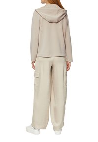 Hellerbeige Kapuzenpullover mit glatter Textur, kombiniert mit locker sitzenden beigen Cargo-Hosen mit Seitentaschen. Weiße Sneakers runden das Outfit ab.