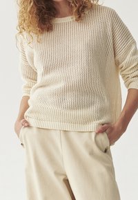 Helles beige Strickpullover mit einem lockeren, atmungsaktiven Design und einem gerippten Saum. Kombiniert mit cremefarbenen Hosen, die eine lässige Passform zeigen.