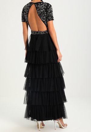 Rückenansicht einer Frau, die ein schwarzes stufiges Tüllkleid mit kurzen Ärmeln und einem perlenbesetzten, offenen Rückenkorsett trägt, kombiniert mit weißen Sandalen mit hohen Absätzen.