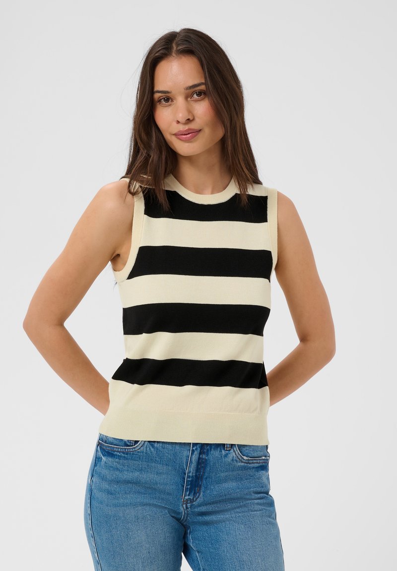 Femme portant un haut sans manches à rayures horizontales noires et blanches et un jean bleu, debout les mains derrière le dos sur fond gris.
