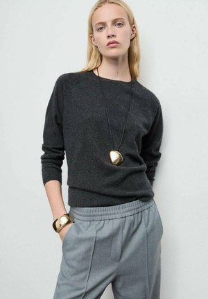 Maglione di lana grigio scuro con scollo rotondo, abbinato a pantaloni grigio chiaro. Accessori includono un braccialetto dorato e una collana con pendente geometrico.