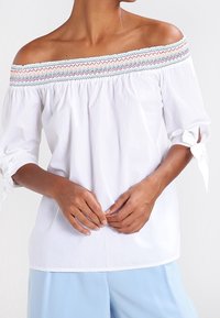 Donna che indossa una blusa bianca off-shoulder con bordi ricamati multicolore e maniche legate, abbinata a pantaloni azzurri.
