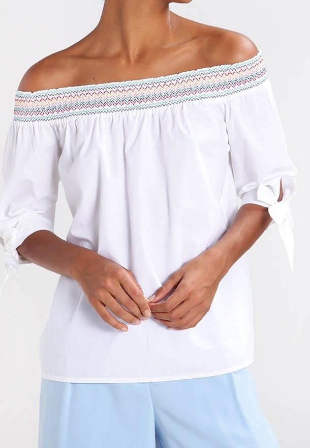 Donna che indossa una blusa bianca off-shoulder con bordi ricamati multicolore e maniche legate, abbinata a pantaloni azzurri.