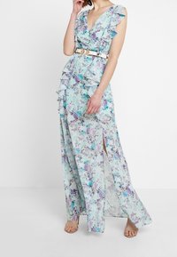 Femme portant une robe maxi florale bleu clair avec des manches volantées, une fente sur le devant et une ceinture blanche, se tenant devant un fond blanc uni.