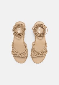 Beige braided leather sandals with circular accent detail, flat sole, and a unique intertwined design. Textured woven straps provide detail.