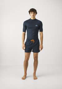 Granatowa koszulka rashguard z krótkim rękawem z logo; dopasowane granatowe szorty kąpielowe z jaskrawoczerwonym sznurkiem; lekki, gładki materiał, minimalistyczny design.