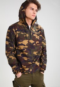 Chaqueta de polilona camuflaje con capucha, que presenta un cierre de media cremallera en la parte delantera, puños elásticos y una mezcla de colores verde, marrón y beige.