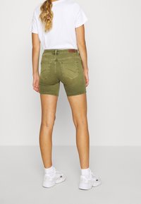 Bermudas de mezclilla verde oliva con un diseño ajustado, cinco bolsillos y bordes rematados, combinadas con una camiseta blanca lisa y zapatillas blancas.