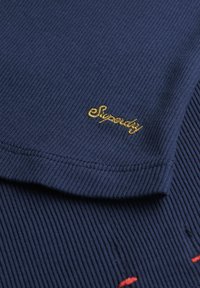 Navyblauwe geribbelde katoenen t-shirt met korte mouwen. Heeft een gouden geborduurd logo "Superdry" op de borst, met rode accenten.